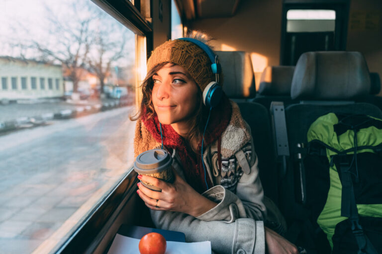 A girl is sitting on a train with headphones on her ears, holding coffee in her hand, looking at the sunset