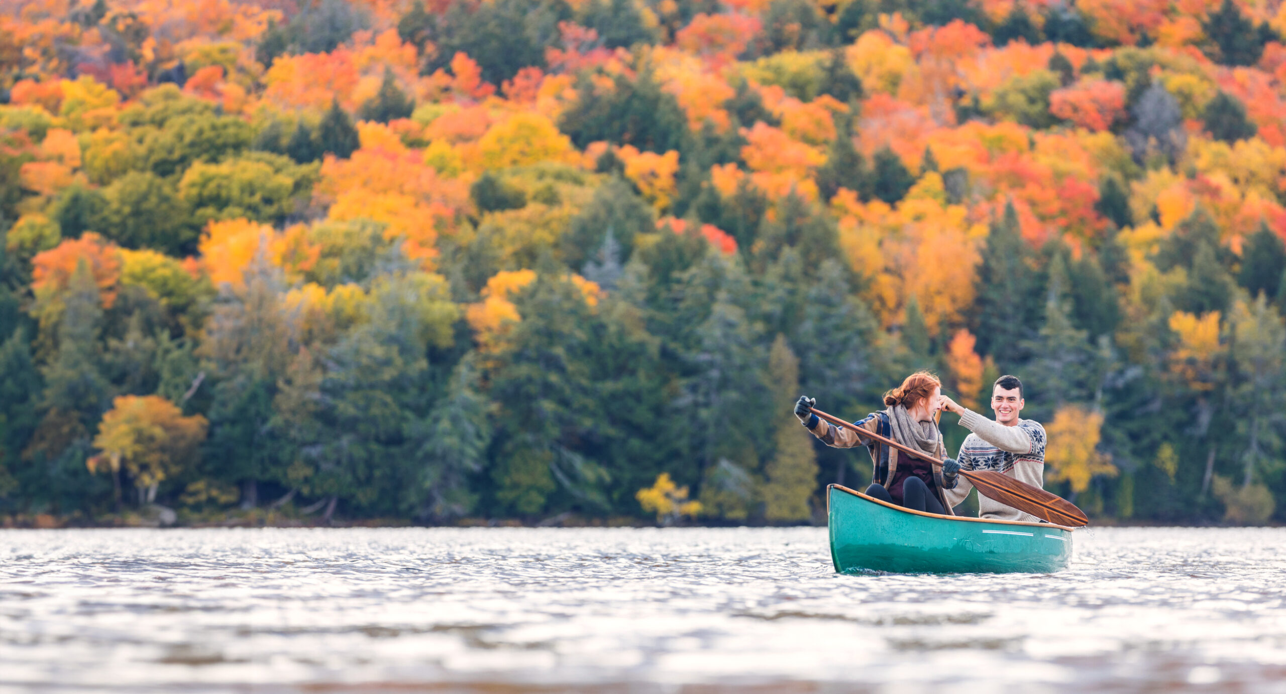 A couple on a lake canoeing together. The trees behind them are brilliant fall colors