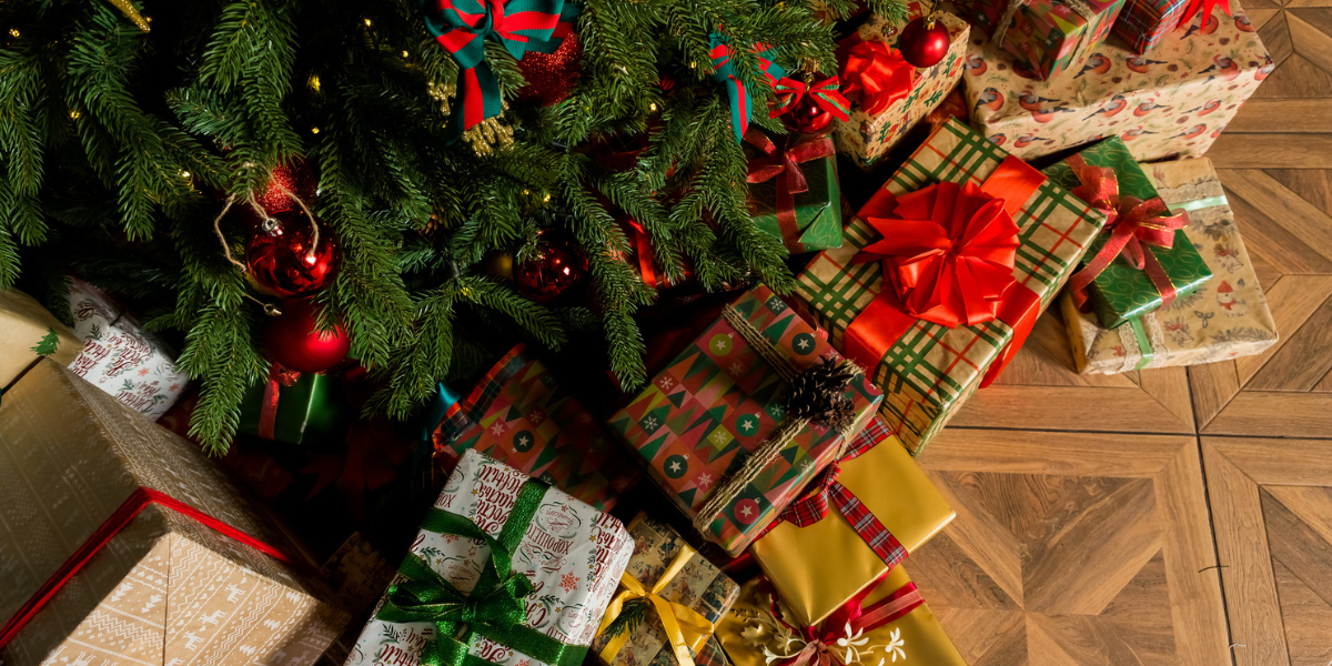 An above-view of gifts under a christmas tree. You see the lower branches of a decorated tree hanging over a variety of festively wrapped presents on a wood floor.