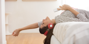 A woman laying backwards on her bed with headphones in as she enjoys music. She is smiling and reach her hand out off the bed and above her head.